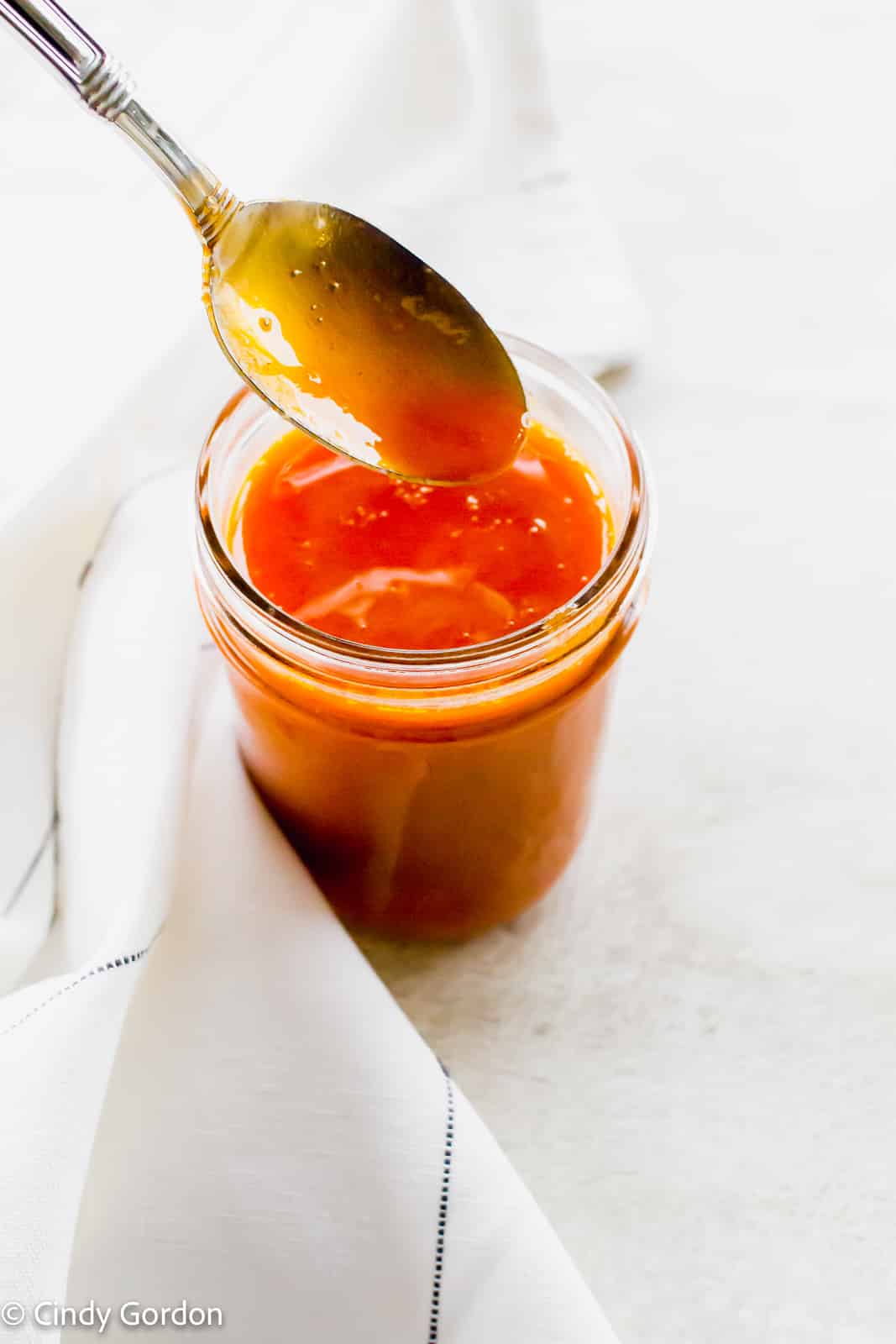 A spoon coated in Buffalo sauce over a glass jar of bright red sauce