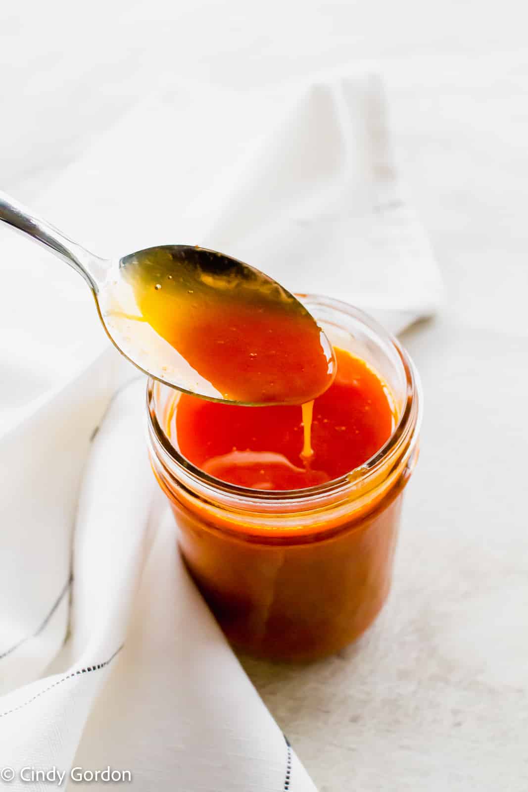 A spoonful of buffalo sauce pouring into a mason jar of more sauce wrapped in a white towel