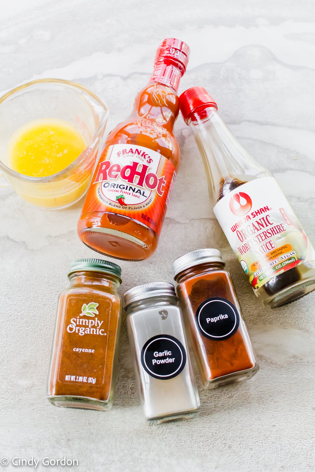 Bottles of hot sauce, vegan Worcestershire sauce, and seasonings with a measuring cup of melted butter