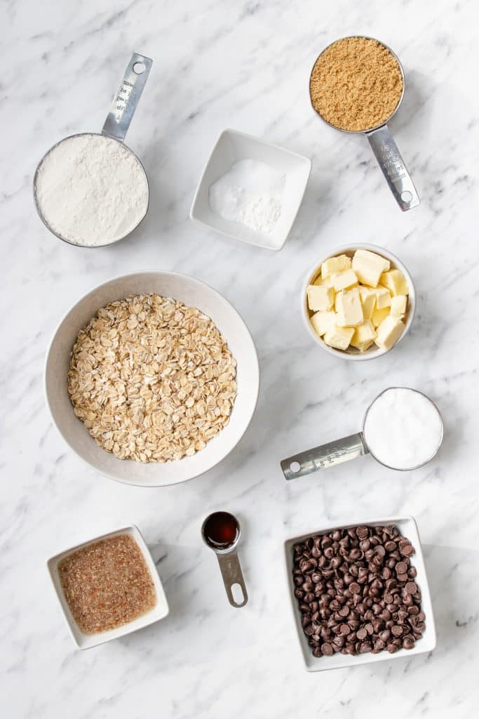 Bowls of butter, rolled oats, sugar, flour, baking soda, brown sugar, and chocolate chips on a granite countertop