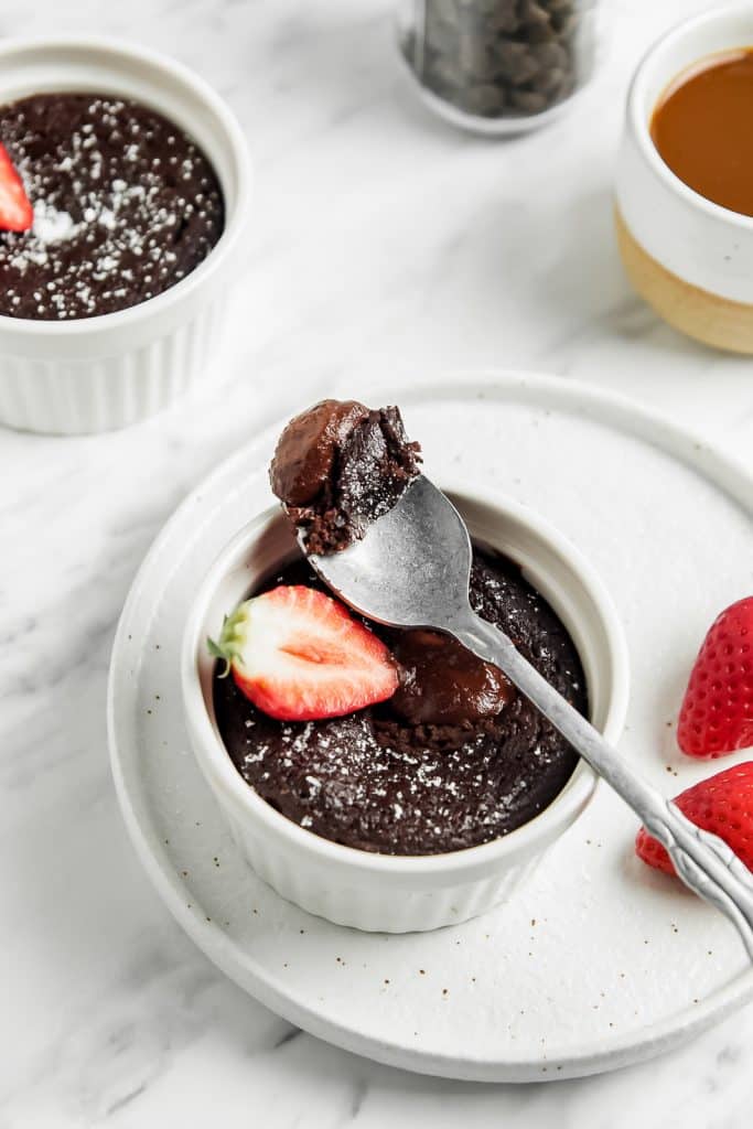 A decadent bite of chocolate mug cake on a spoon over the ramekin of cake with a strawberry slice