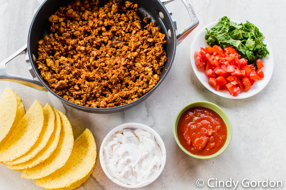 A skillet of crumbled tofu surrounded by hard taco shells, sour cream, salsa, lettuce, and diced tomatoes
