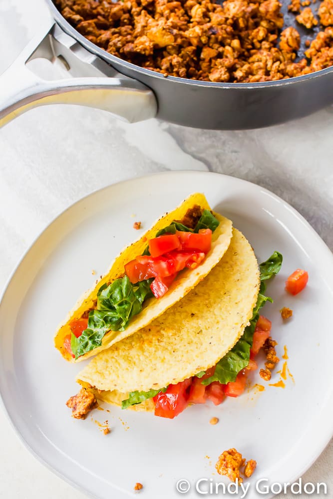 Hard taco shells stuffed with tomatoes, lettuce, and crumbled tofu on a white plate