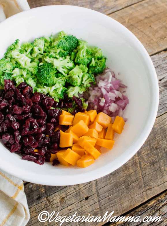 ingredients for broccoli salad in a bowl, including dried cranberries, yellow cheddar cheese, broccoli florets, and diced red onion.