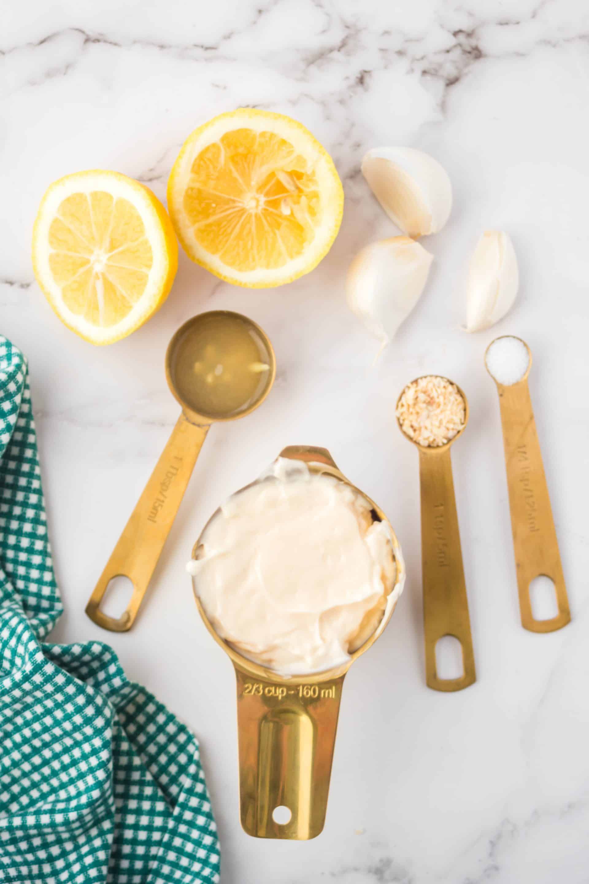 measuring cups and spoons of salt, lemon juice, minced garlic, and mayonnaise on a granite countertop