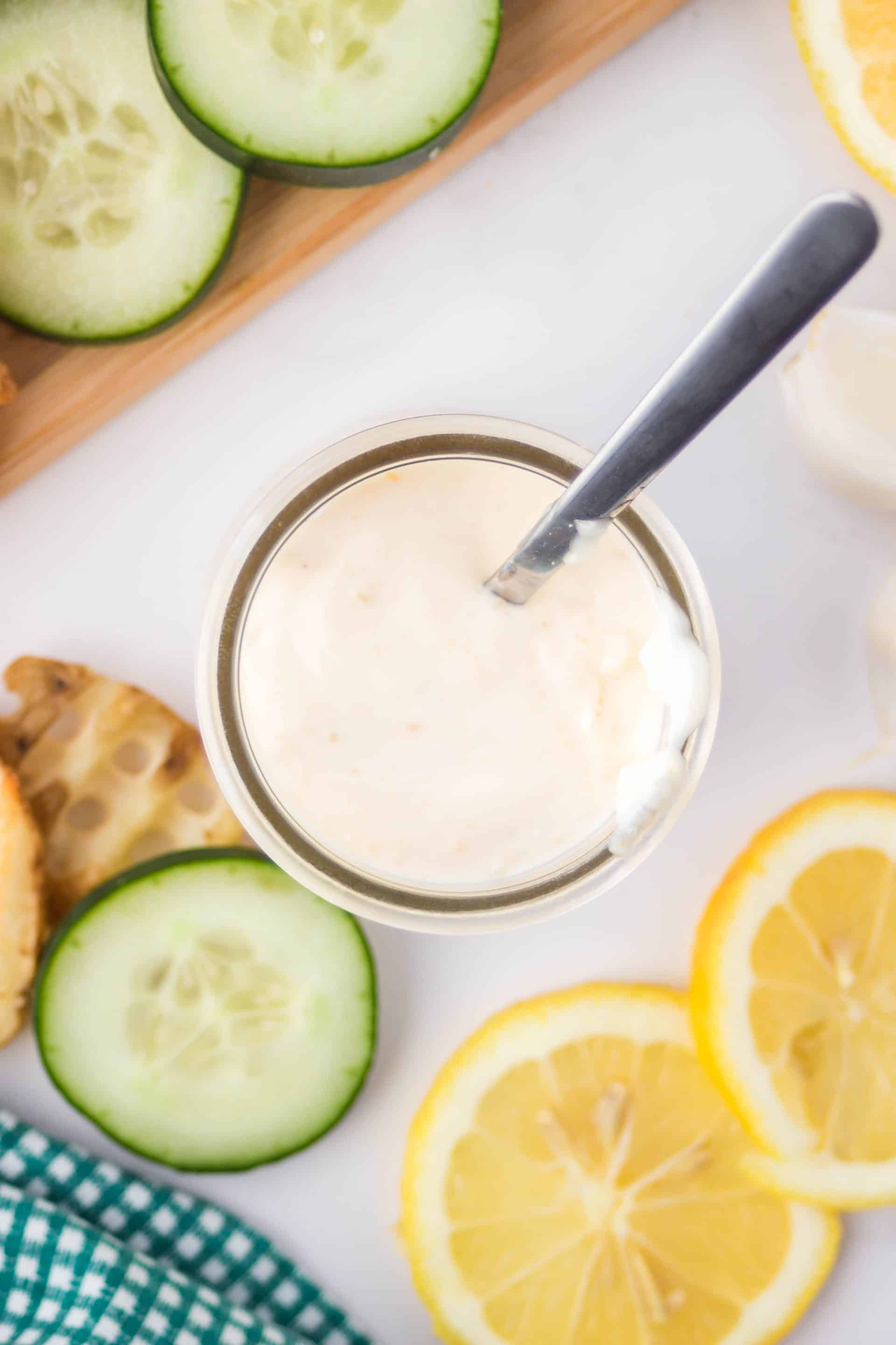 Overhead shot of vegan aioli in a glass jar on a white countertop with lemons and cucumbers