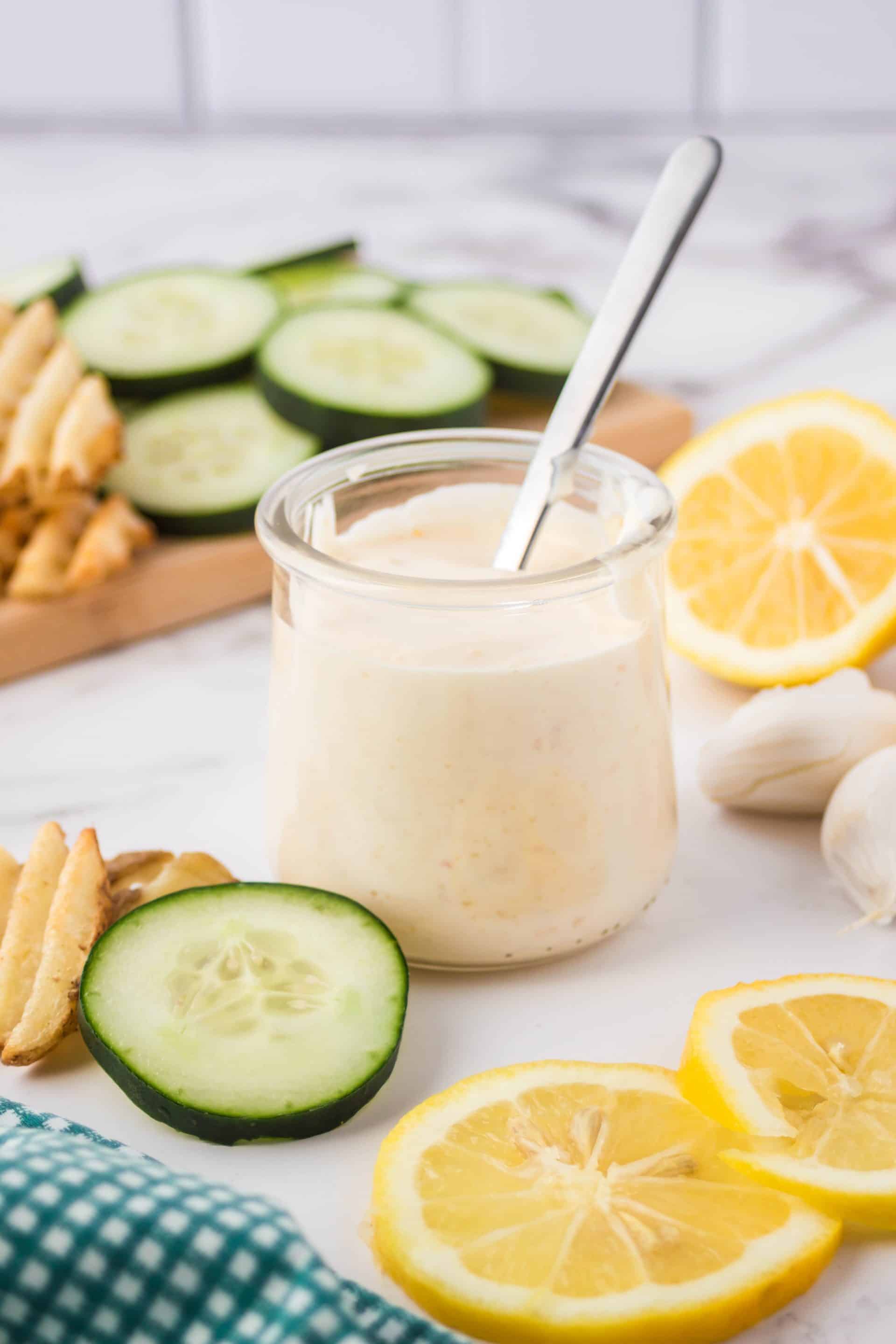 A clear jar of vegan aioli sauce with a spoon in it surrounded by cucumbers, garlic, and lemons