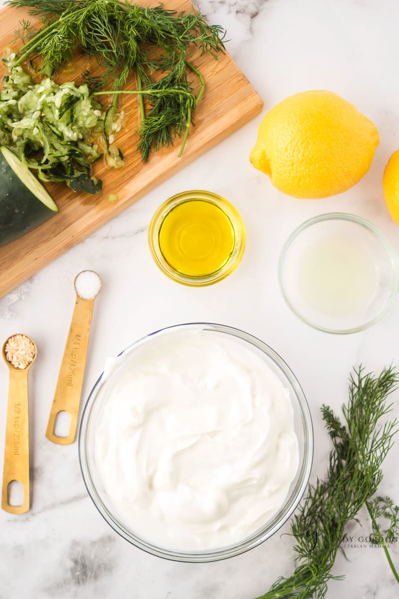 Bowls of Greek yogurt, lemon juice, olive oil, salt and minced garlic next to lemons and fresh dill