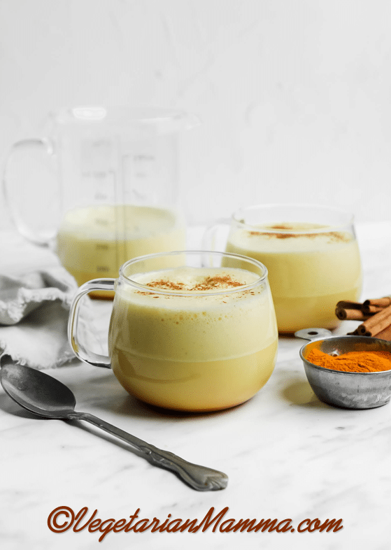 vertical picture of turmeric coffee and turmeric latte in clear mug with tarnished spoon in front. A clear container of the mixtures behind it to the left.
