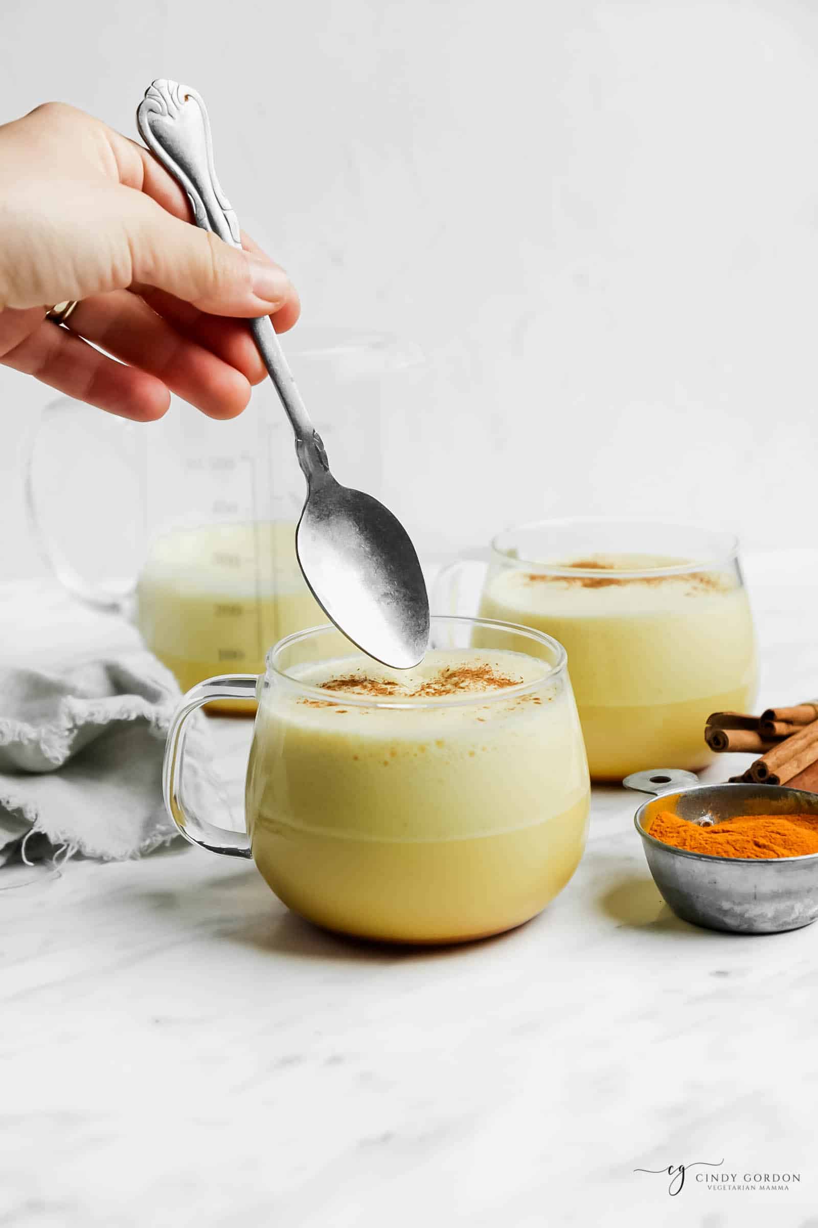 a hand holding a spoon above a clear mug of turmeric latte