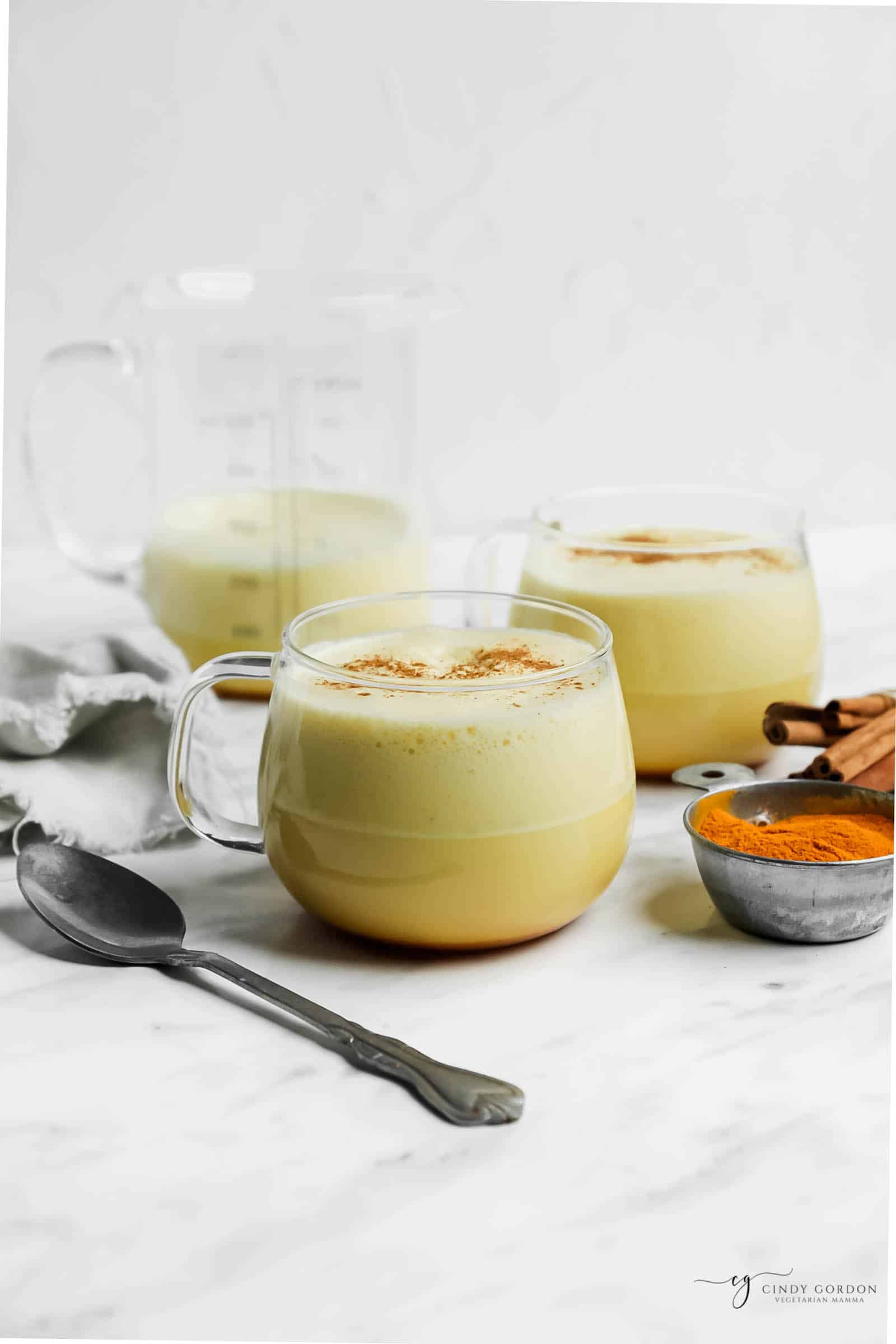 vertical picture of turmeric coffee and turmeric latte in clear mug with tarnished spoon in front. A clear container of the mixtures behind it to the left.