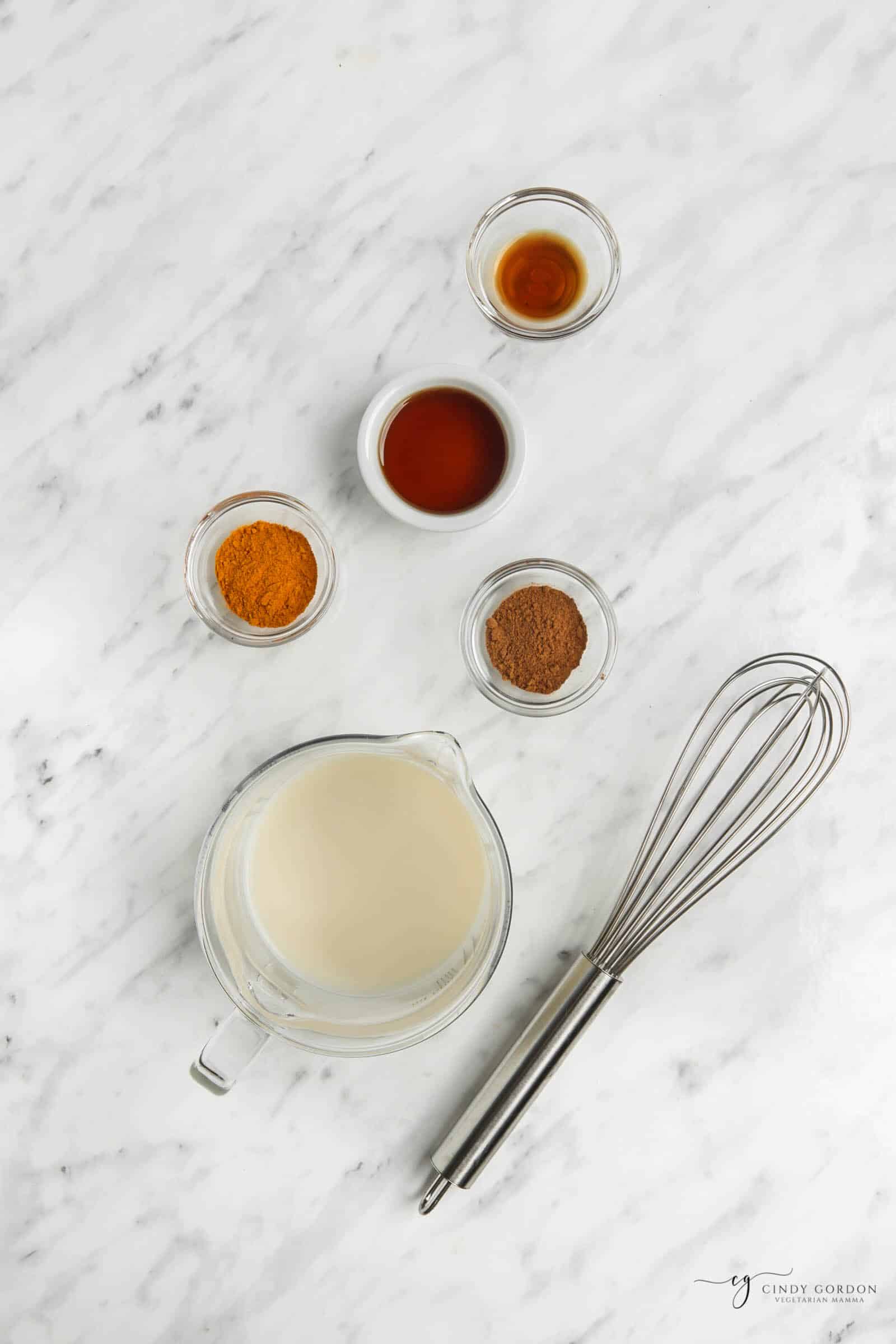 measured ingredients for turmeric latte. Ingredients are in clear and metal containers on a white marble background.