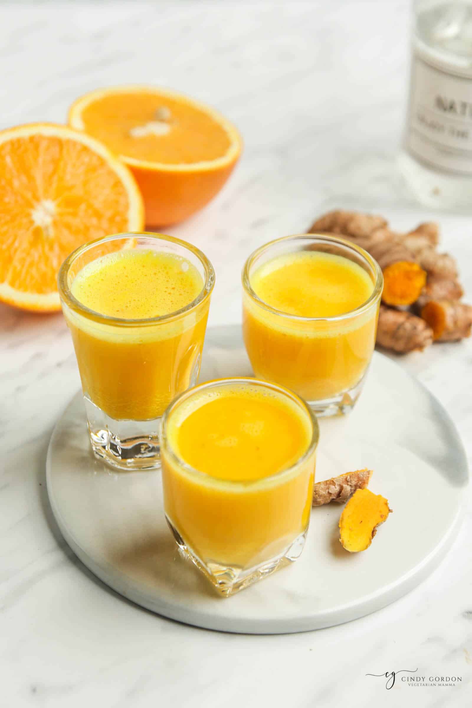 Vertical photo angled downward, showing three clear glasses filled with an orange liquid on a white trivet. Broken pieces of fresh turmeric root beside the glasses.