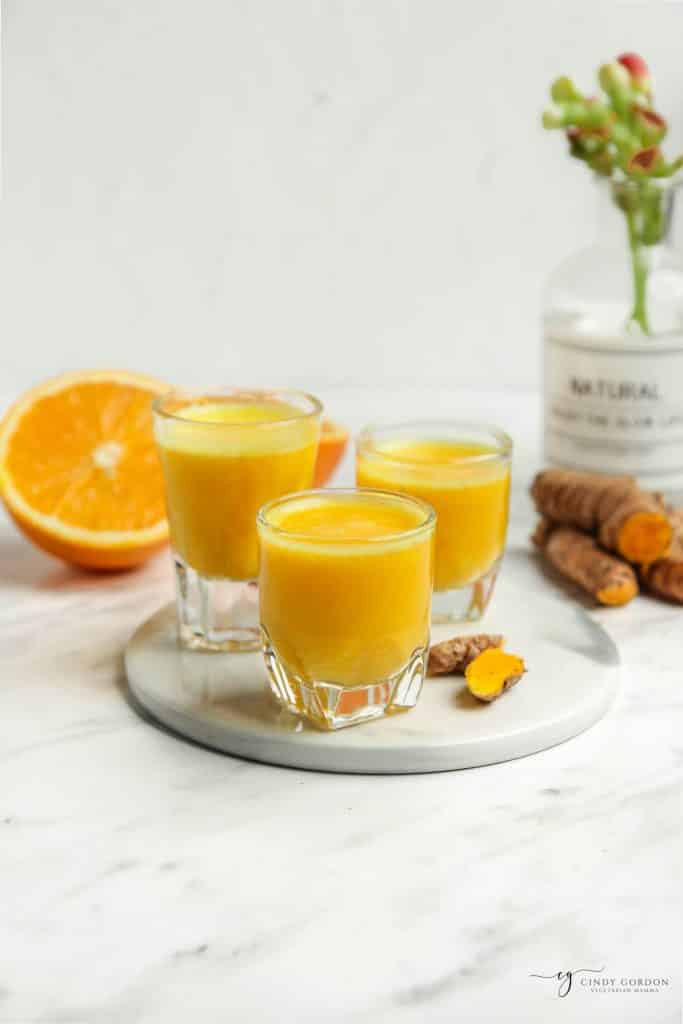 vertical angled shot of orange liquid in three glasses, with orange half to the left, fresh turmeric to the right