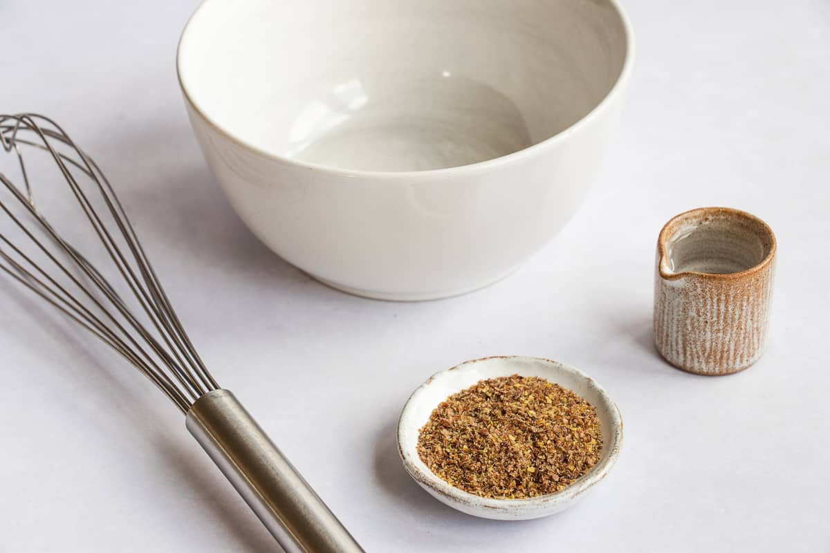 A whisk, a white mixing bowl, a small pitcher of water, and a bowl of ground flaxseed