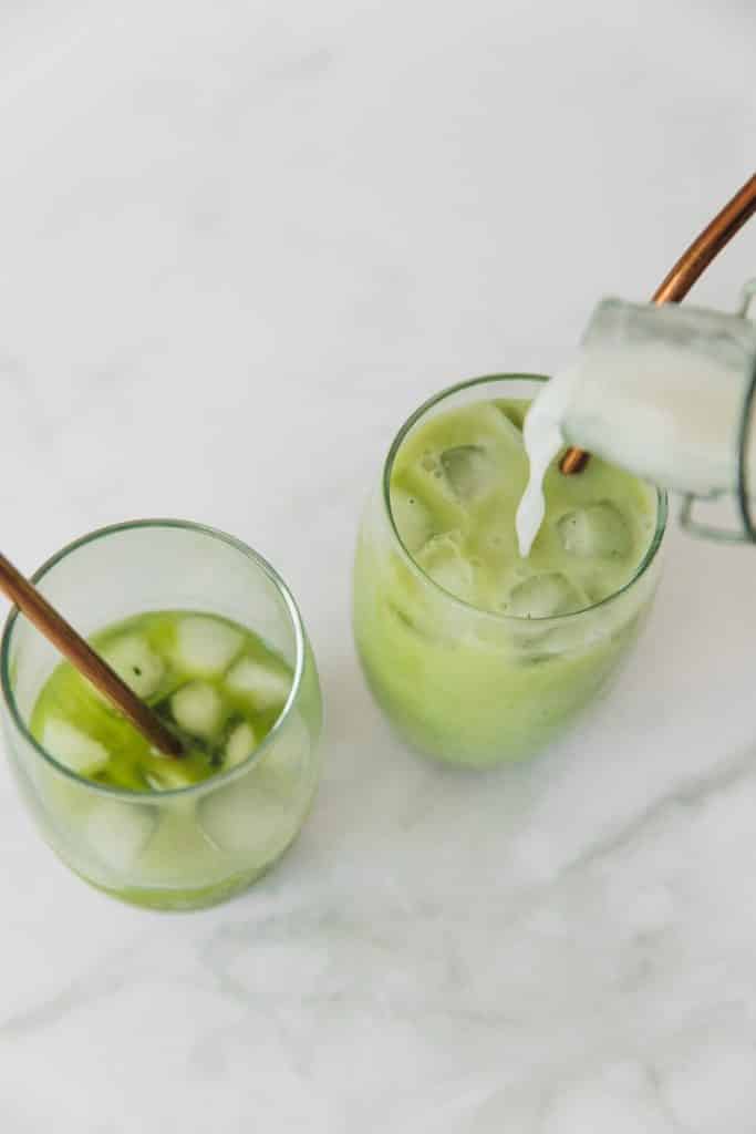 two clear glasses with ice. One filled half way with green liquid. One is being filled up to the top with a white liquid being poured into a green liquid.