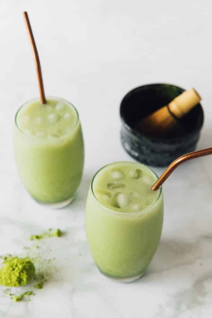two clear glasses filled with ice and mint green liquid. Matcha mixing bowl with mini broom to the top right.