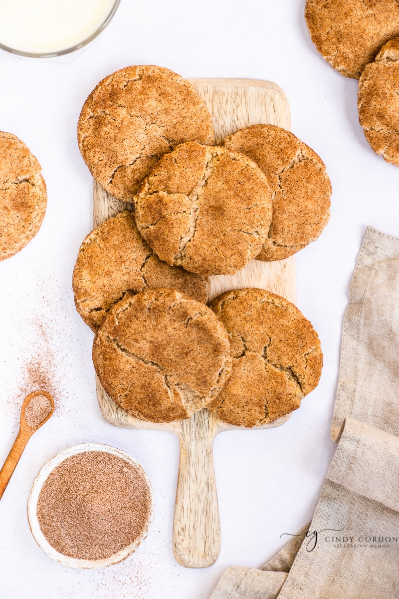 Six large snickerdoodle cookies on a wooden cutting board surrounded by more cookies and a towel