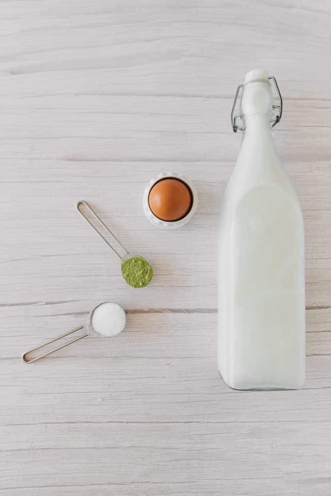 vertical photo. Brown egg, white liquid in a glass bottle, green powder in a measuring spoon and white granulated powder in a measuring spoon.