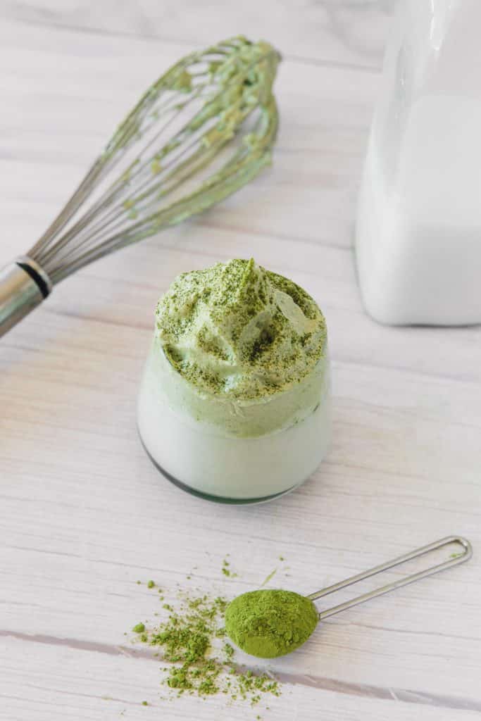 vertical image with a clear glass filled with white liquid and creamy green topping. Top of the picture is a whisk and bottom of the picture is a measuring spoon with green powder