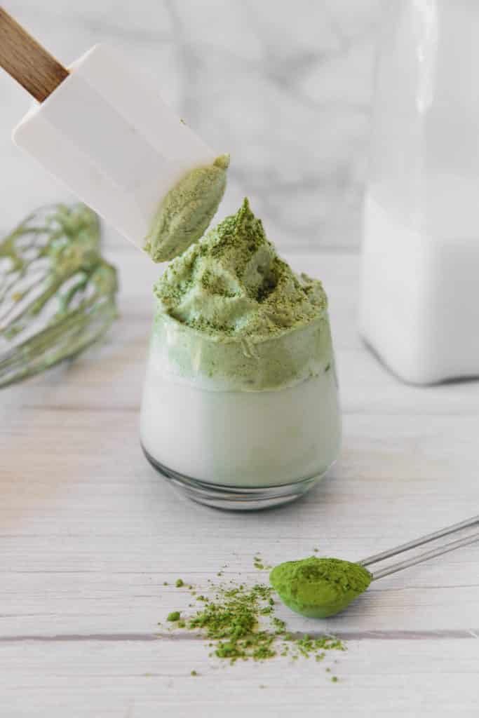 clear glass filled with white liquid and a spatula dolloping green cream on top. Measuring spoon with green powder in front of the glass.