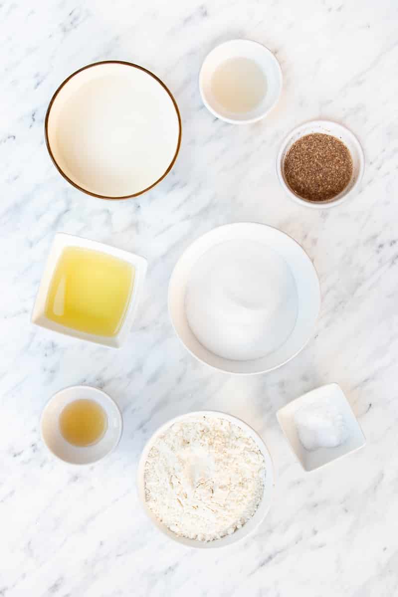 Bowls of sugar, cinnamon, flour, butter, vanilla extract, almond milk, vinegar, and salt on a countertop