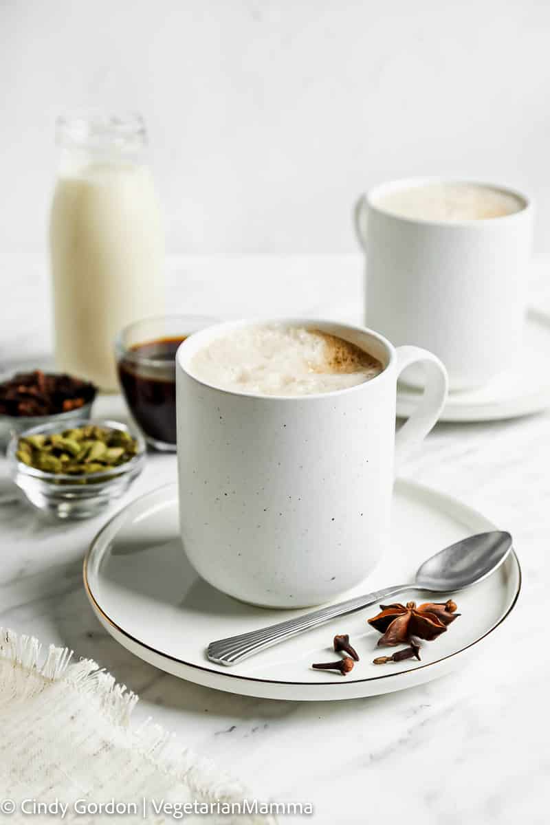 white foamy liquid in white mug on a white plate with a spoon and spices on top. In the back are clear small bowls of spices and maple syrup and a glass jar of milk. 