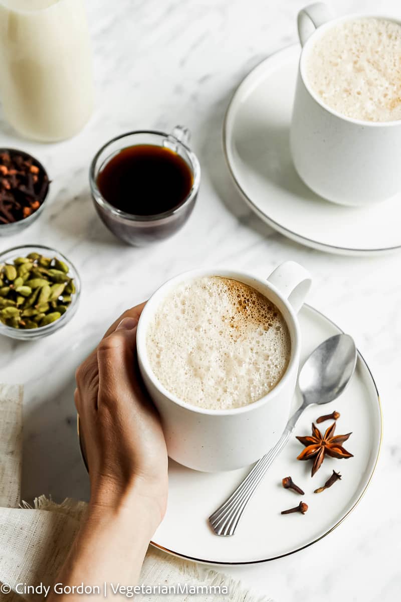 foamy creamy liquid in white cup on a white plate with a handing holding the cup from the left. On white plate is spices and a silver spoon. In the upper right hand corner is another white up filled with foam on a white plate.