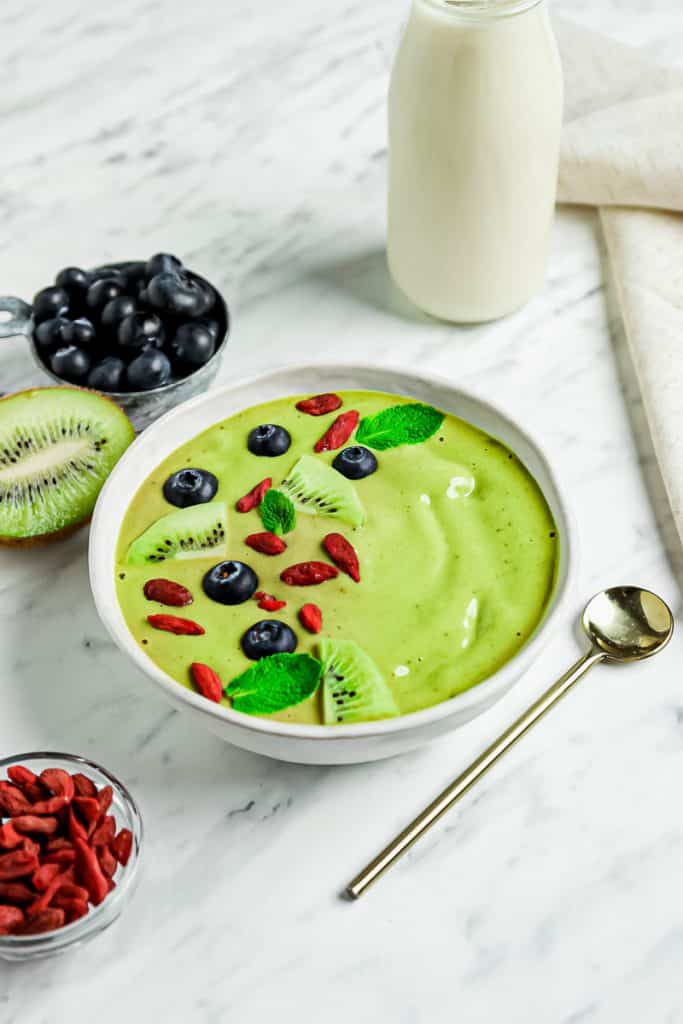 Angled down vertical photo. bottom left has a clear glass bowl of red goji berries. A golden spoon with a long slender handle to the bottom right. Middle has white bowl will creamy green liquid topped with blueberries, mint leaves, kiwi pieces and goji berries. Pictured to the left is a blueberries and kiwi. A glass container of milk to the top right.