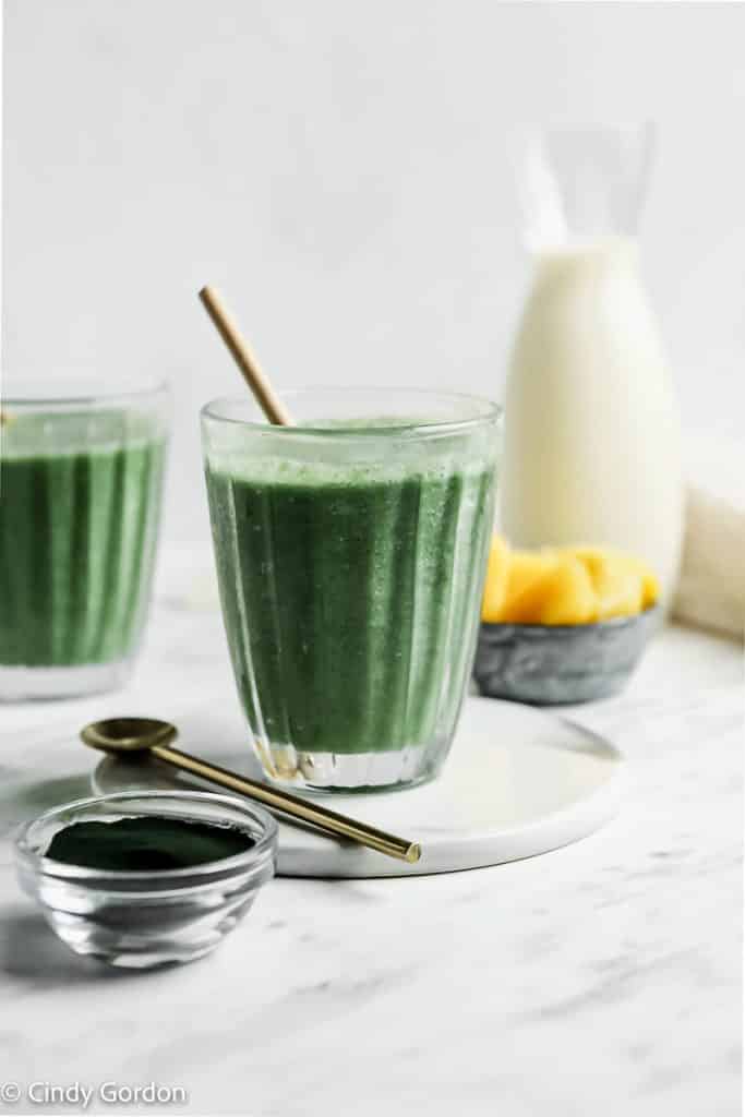 dark green smoothie in clear glass with a gold spoon on a white plate. Diced pineapple chunks in the back ground. Spirulina in glass bowl.