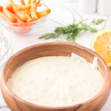 a wooden bowl filled with creamy tartar sauce in front of a bowl of carrots, a bunch of herbs, and half a lemon