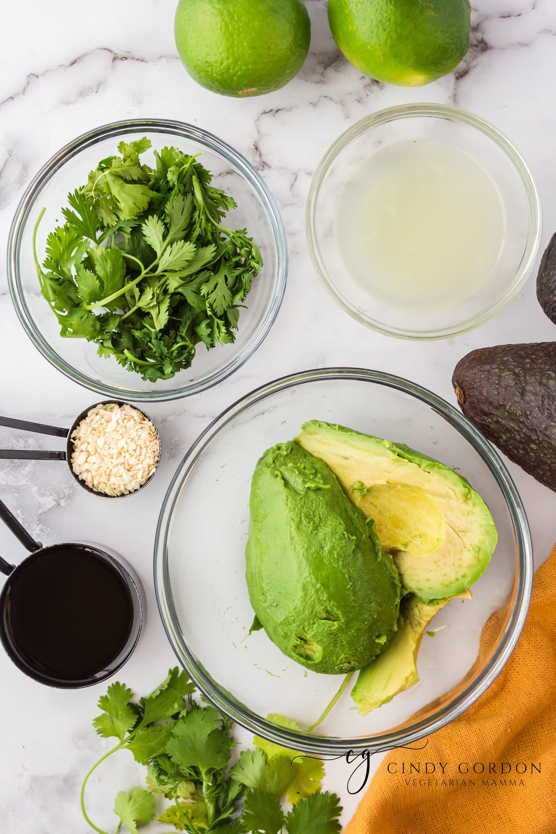 Ingredients for vegan avocado dressing, each in separate bowls on top of a marble counter with an orange towel on the side.