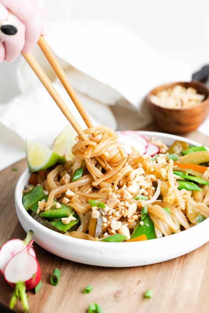 a shallow white bowl filled with vegetable pad thai being eaten with chopsticks. The bowl is on a light wooden cutting board. 