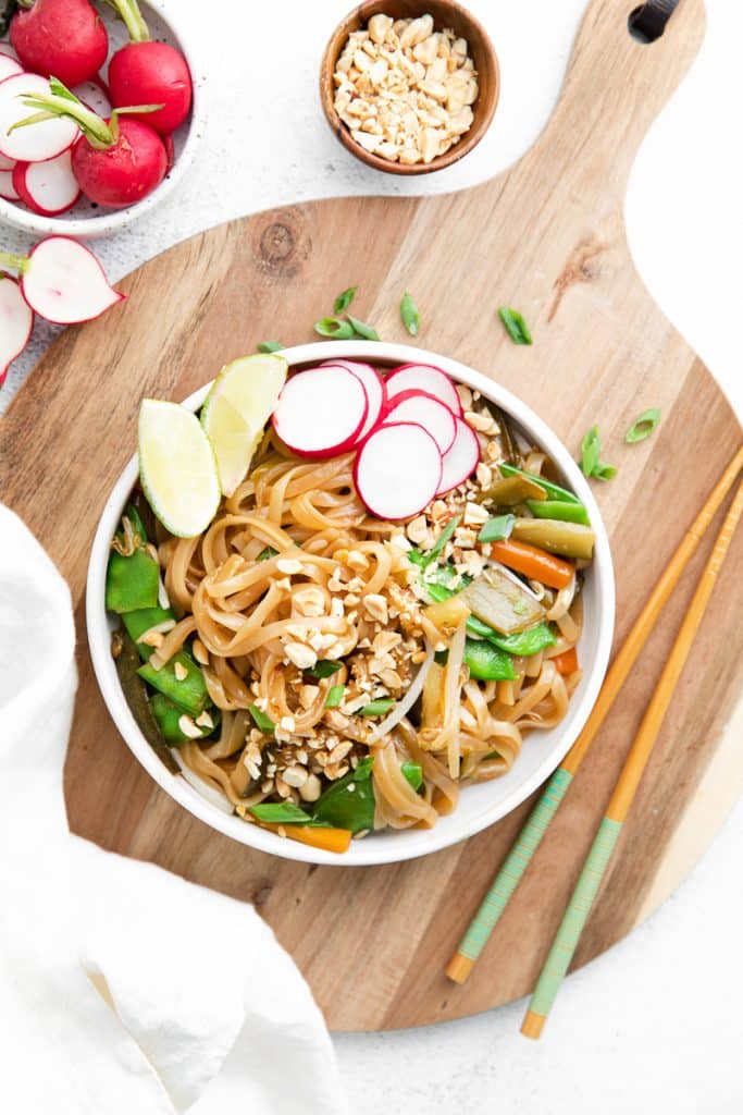 top down view of a wide white bowl filled with pad thai. Garnishes include lime, green onion, peanut, and radishes. The bowl is on top of a wooden cutting board and next to a pair of wooden chopsticks.