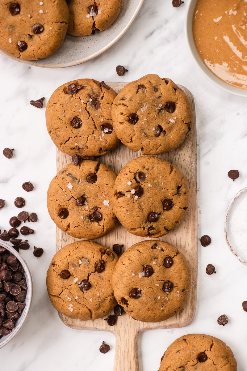 Six peanut butter chocolate chip cookies garnished with sea salt on a wooden cutting board