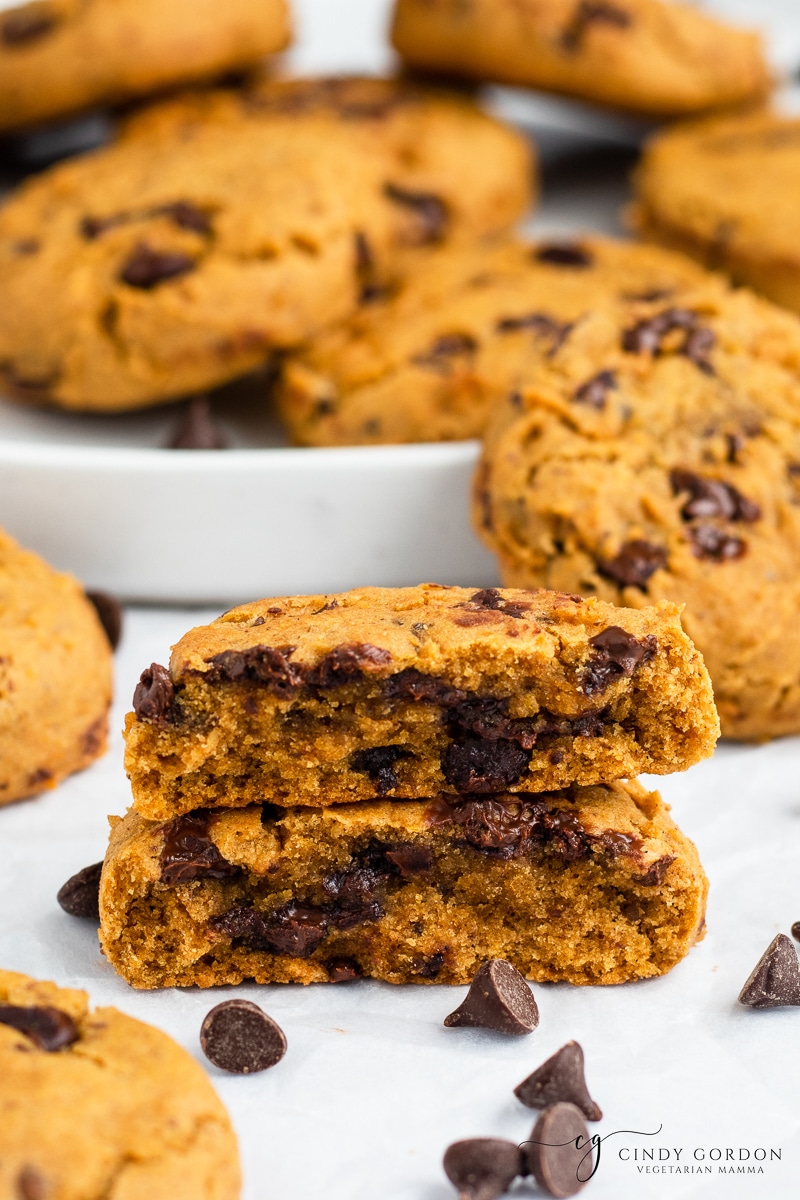 A pumpkin chocolate chip cookie broken in half surrounded by more cookies and chocolate chips