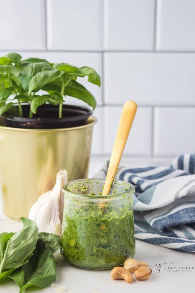 a small jar of green pesto with a gold spoon in it, sitting in front of a potted basil plant