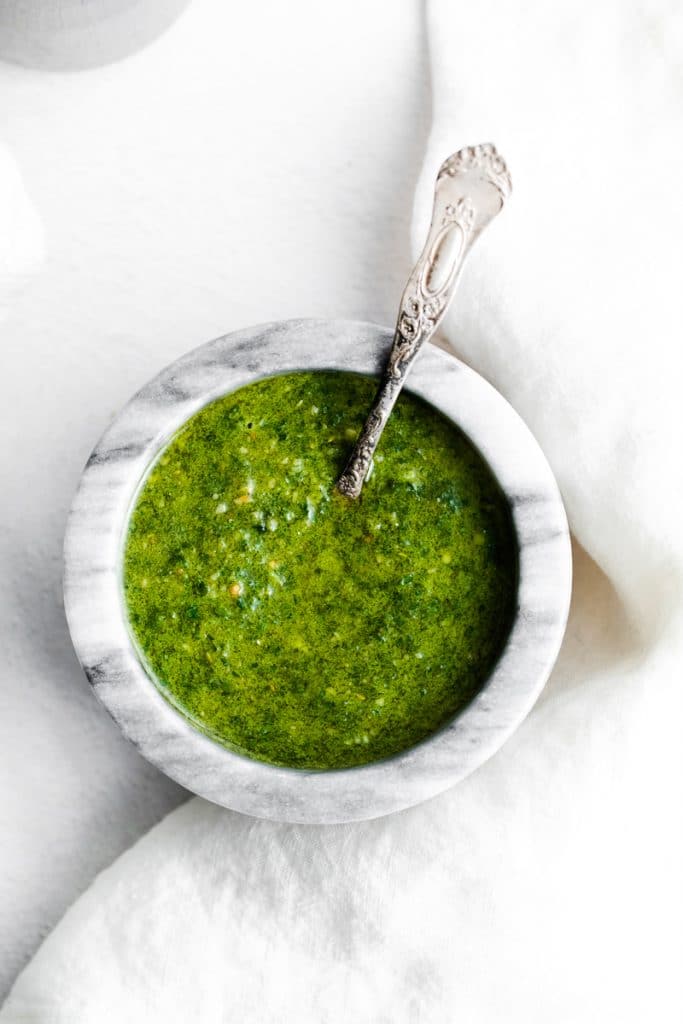 a marble bowl filled with green basil pesto with a silver spoon in it