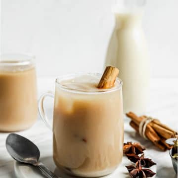 a glass mug filled with an ice chai latte. There is a cinnamon stick in the mug and a glass milk bottle behind it.