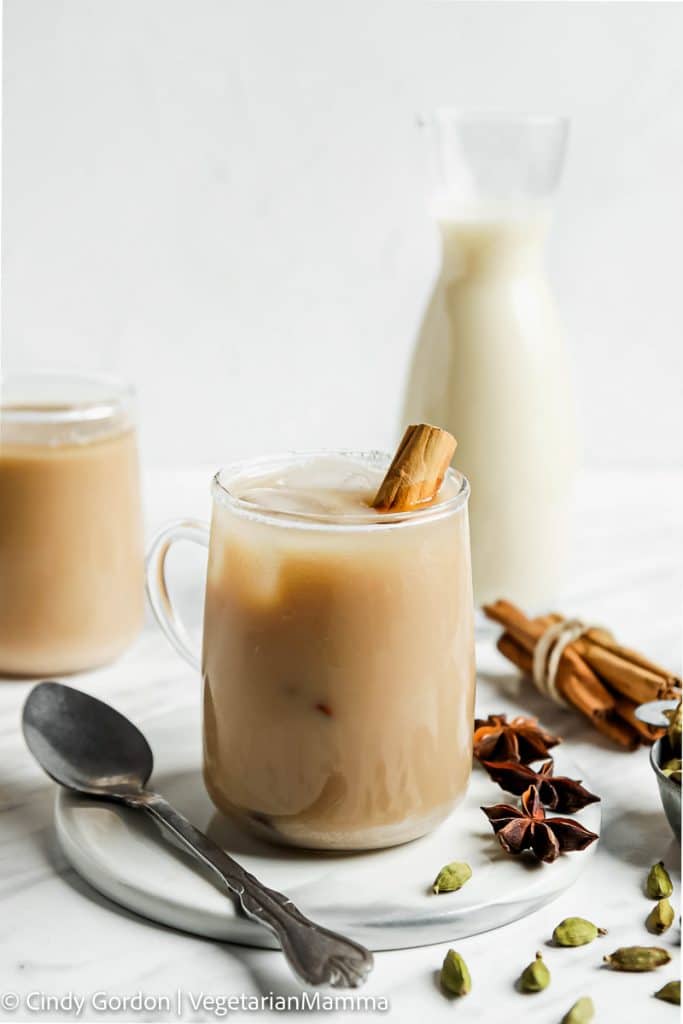 a glass mug filled with an ice chai latte. There is a cinnamon stick in the mug and a glass milk bottle behind it. 