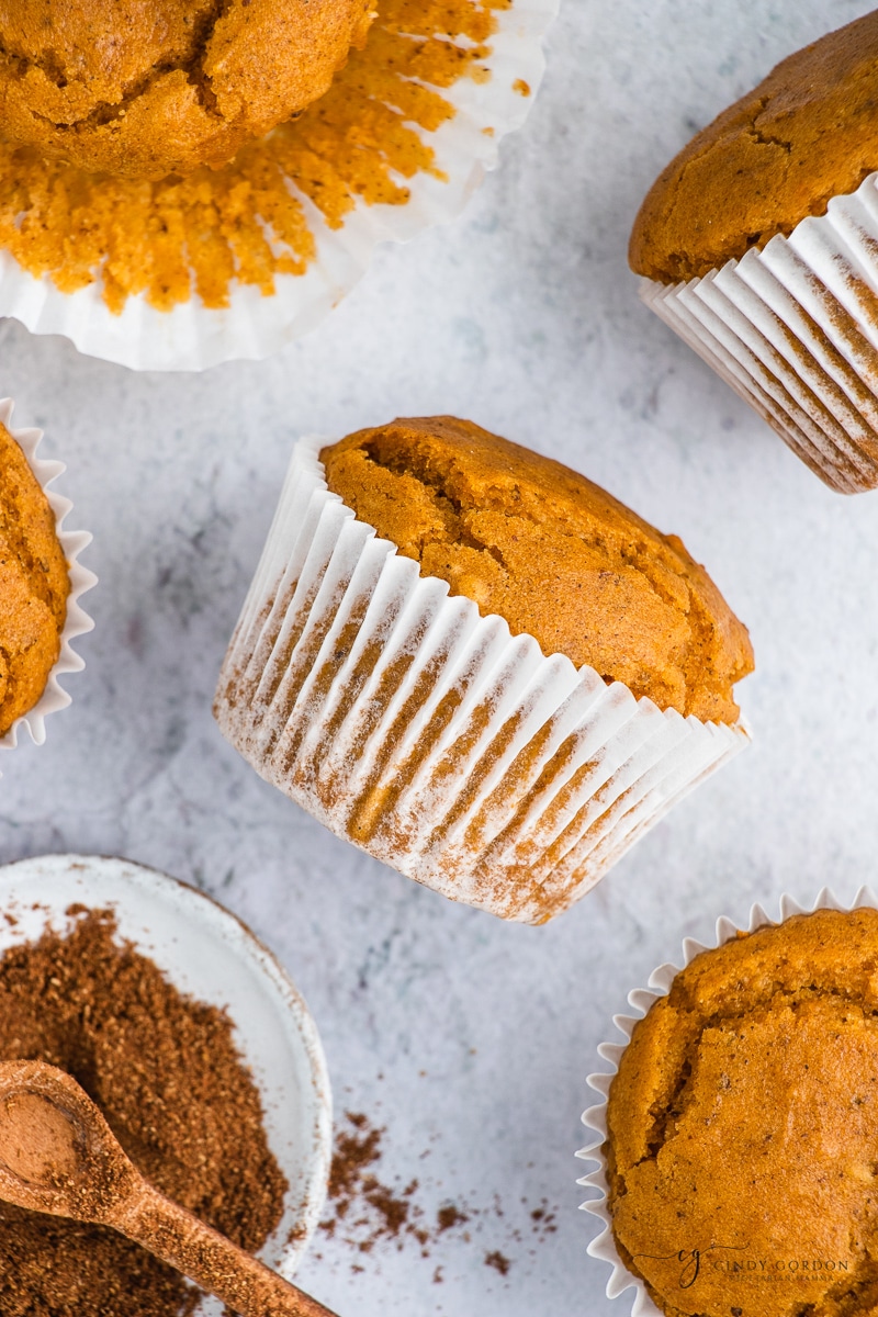 A pumpkin muffin in a white wrapper on its side surrounded by more vegan muffins