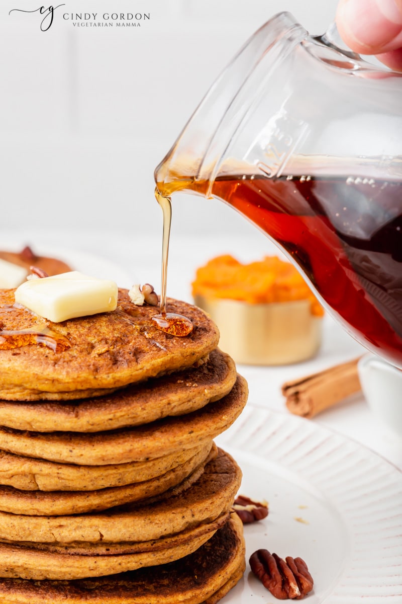 A hand pouring maple syrup over a stack of vegan pumpkin pancakes