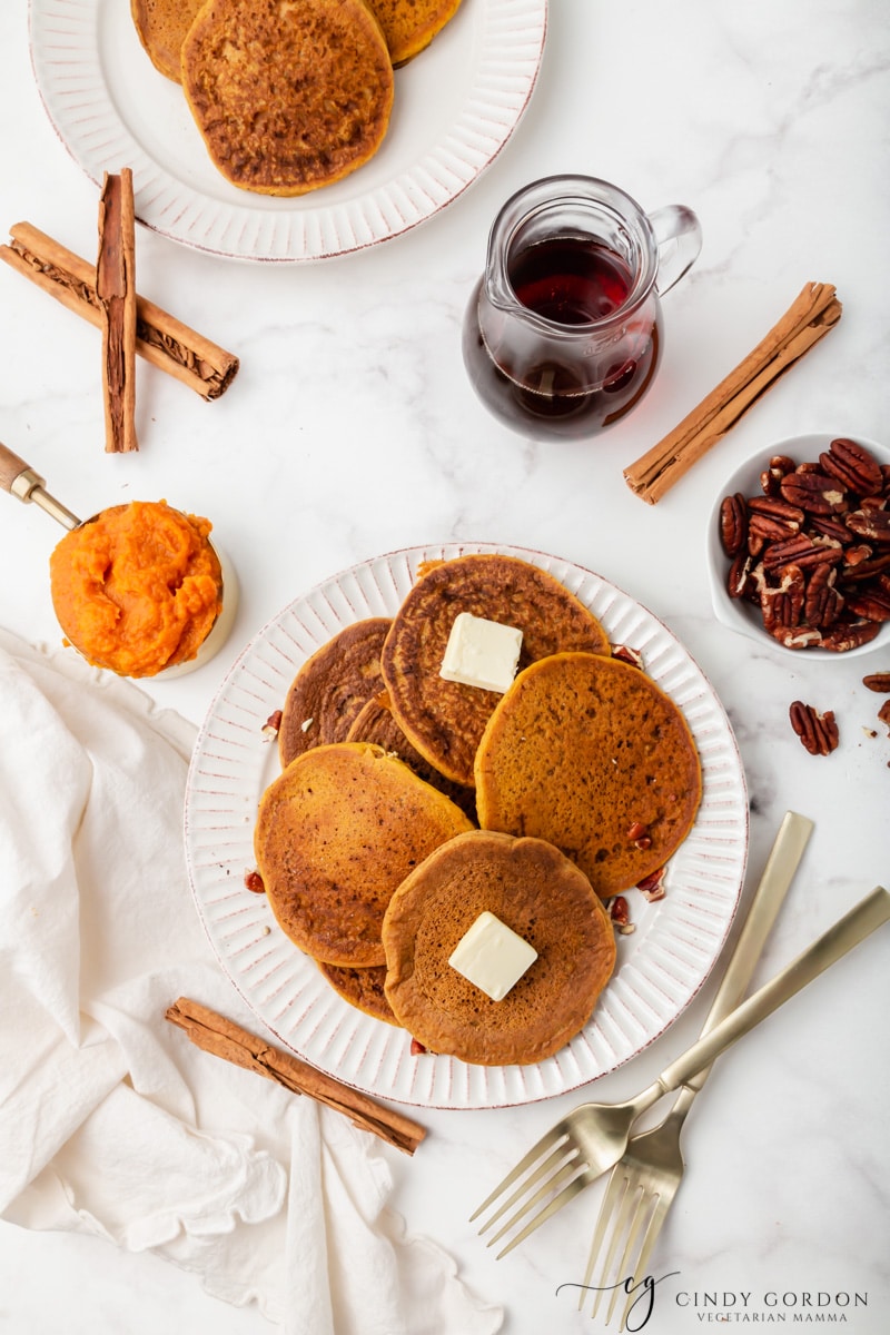 Overhead shot of a plate of pumpkin pancakes surrounded by syrup, pumpkin puree, cinnamon sticks, and pecans