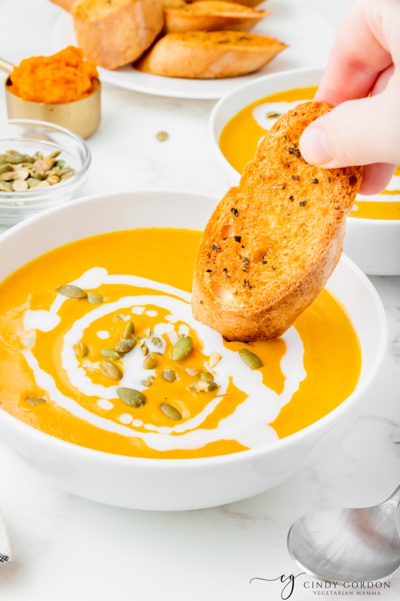 A hand dipping crunchy bread into a white bowl of pumpkin soup