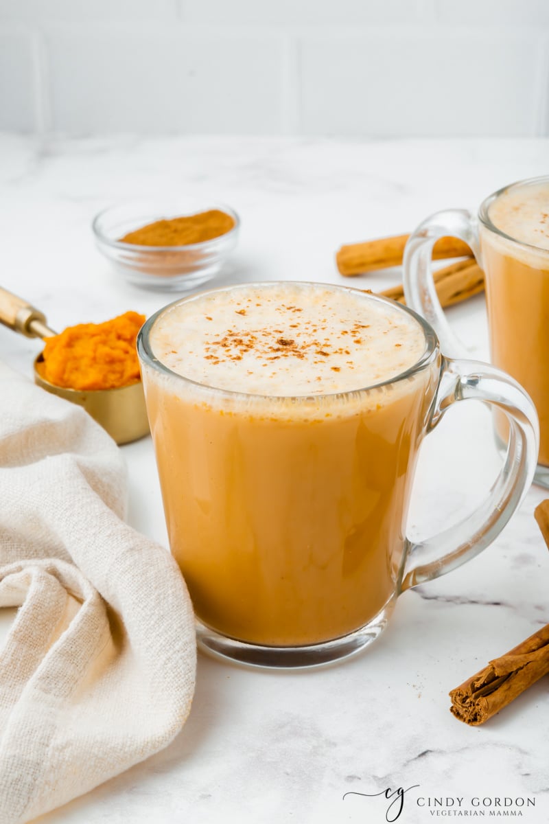 A frothy pumpkin spiced latte in a clear coffee mug surrounded by cinnamon and pumpkin puree