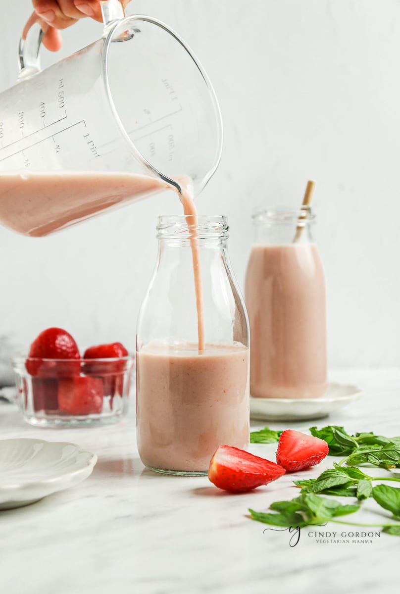 A hand pouring a glass pitcher of strawberry almond milk into a glass bottle