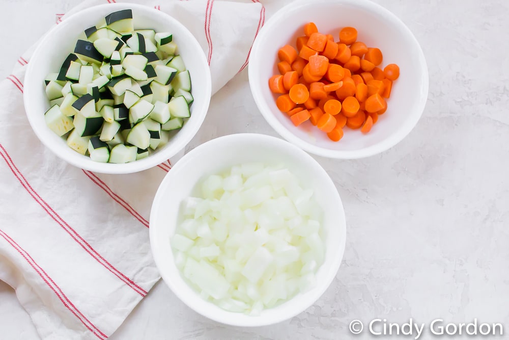 three white bowls, one filled with diced onion, one with diced carrots, and one with diced zucchini
