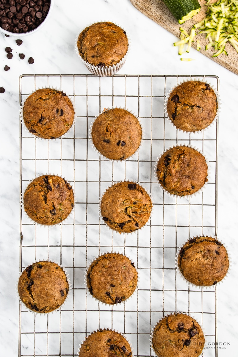 A dozen zucchini muffins on a wire cooling rack surrounded by chocolate chips and grated zucchini