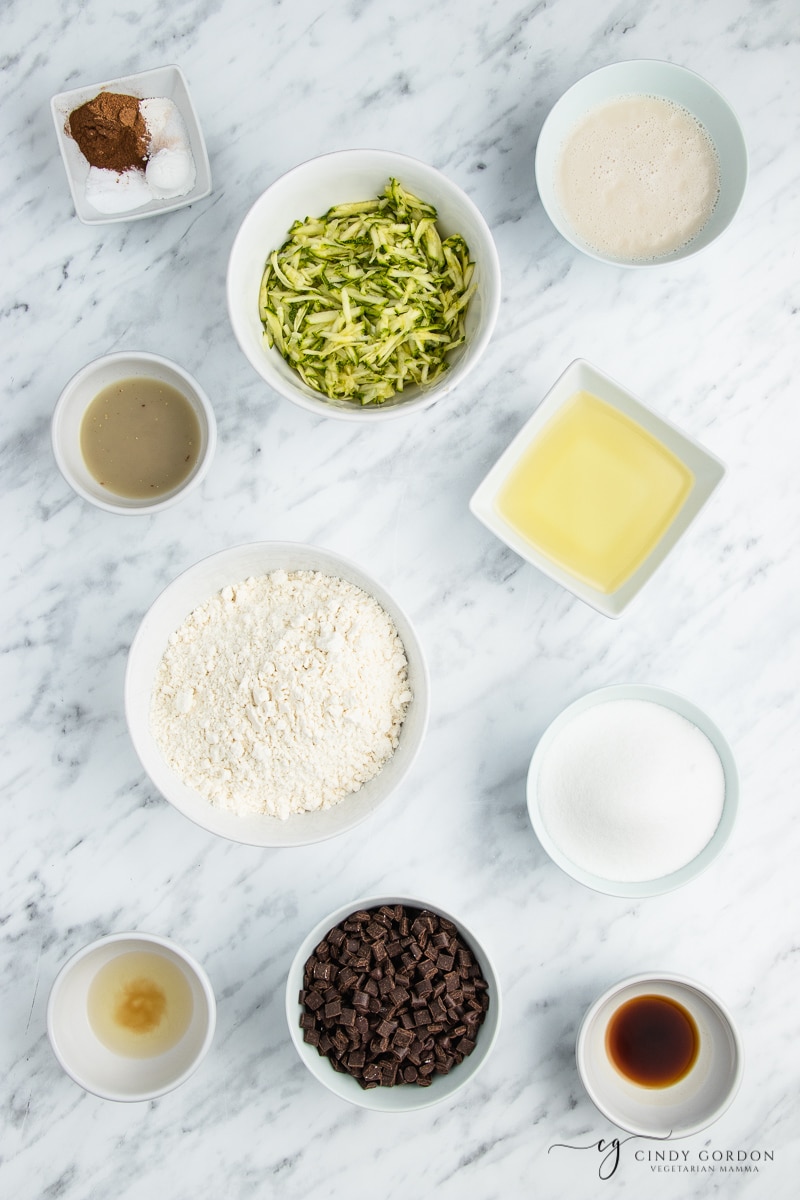 Bowls of grated zucchini, gluten-free flour, chocolate chips, vanilla extract, flax egg, sugar, and oil