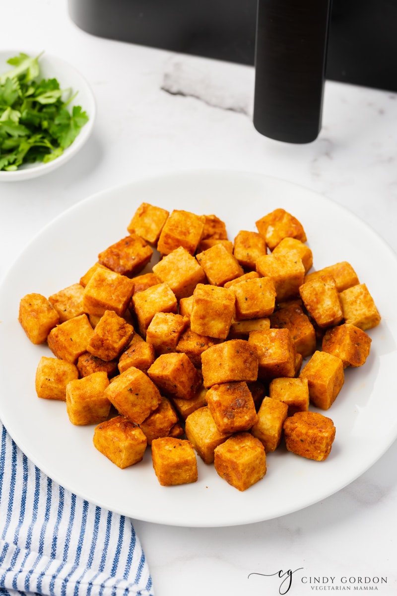 air fryer tofu on white plate on white marble background with green stuff in bowl in top left and black air fryer basket with handle in top right